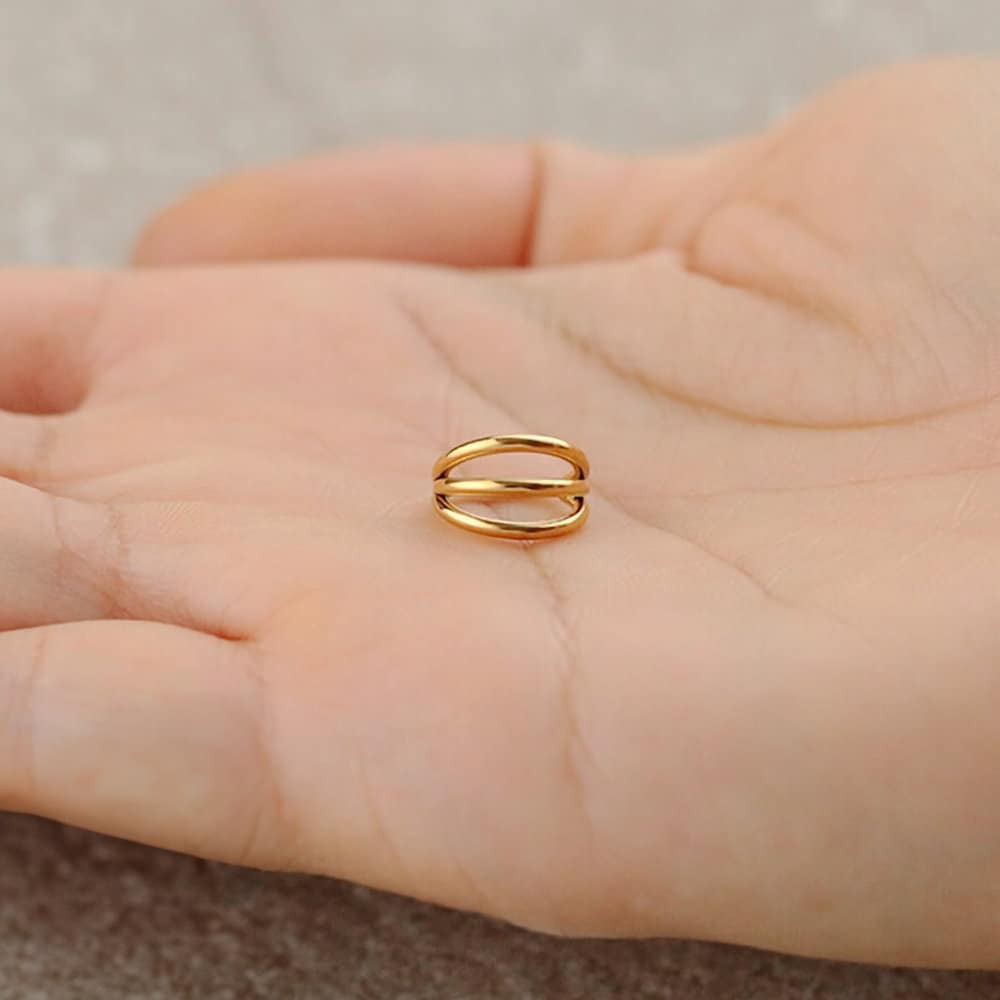 A close-up image of a person holding the Minimalist Gold PVD 316L Steel Septum "Trion" by Kaya Movement, designed with a sleek triple ring effect and subtle elegance. The hand is centered while the shiny surface of the 1.2mm, 8mm diameter ring reflects light against an out-of-focus greyish textured background.